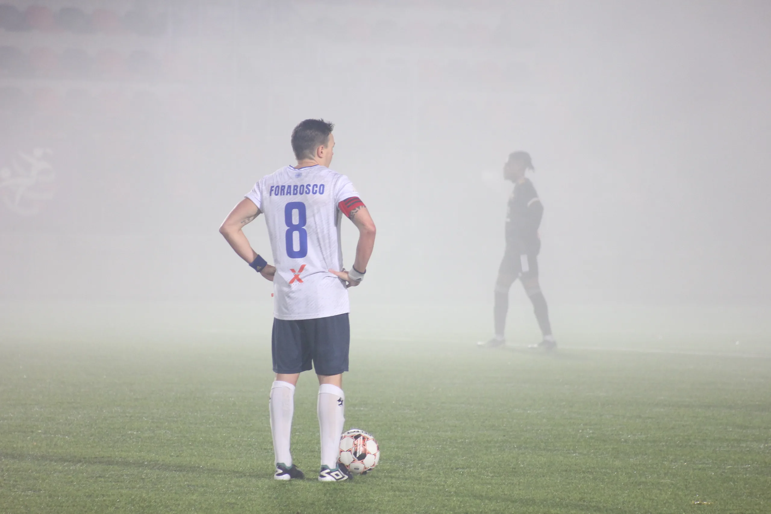 Enrico Forabosco of Krakow Dragoons FC preparing to take a free kick with heavy mist and a blurred Vinsky FC player in the background