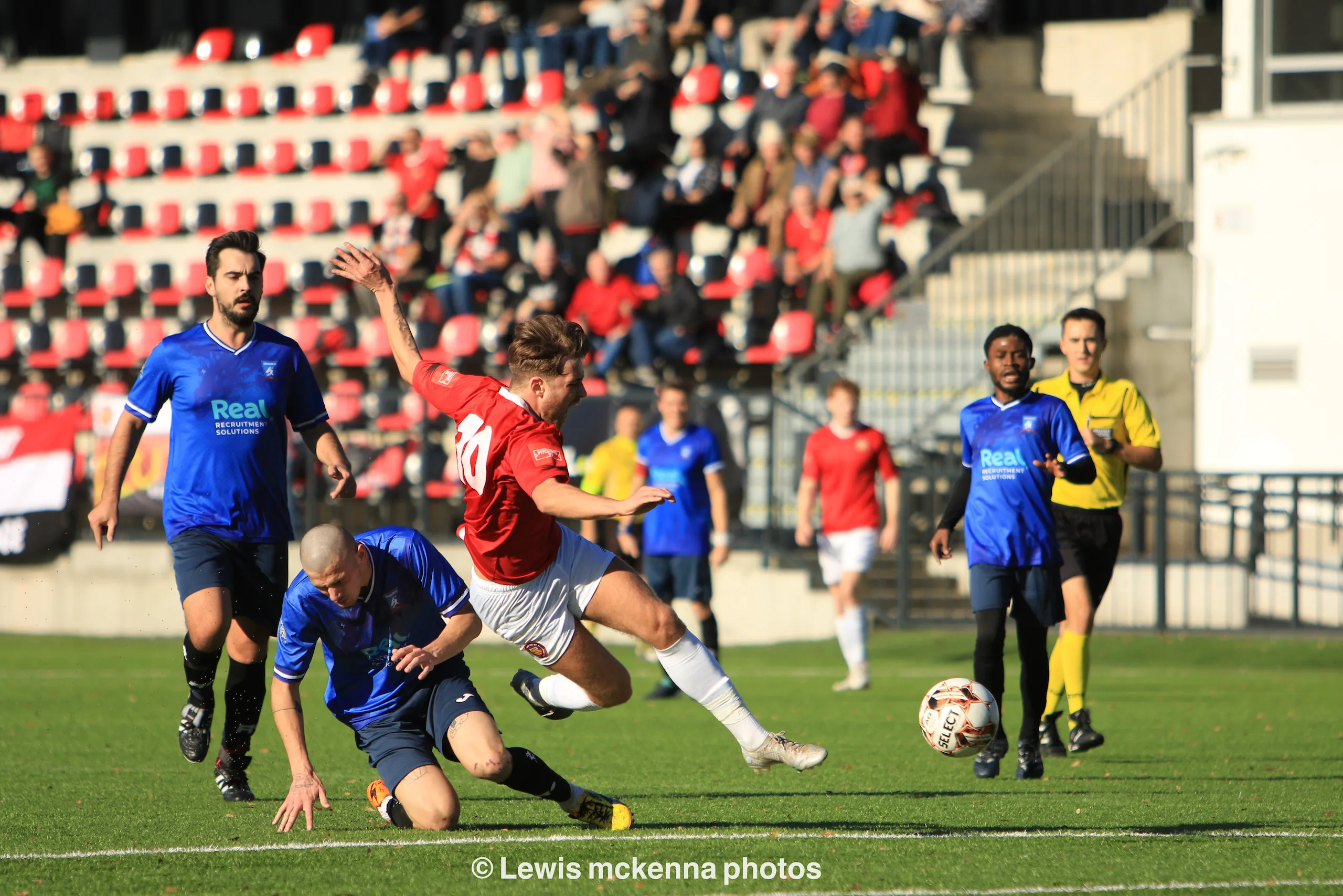 Sasha Arshakian of Krakow Dragoons FC tackles a FC United of Manchester opponent, with other players and the crowd in the background