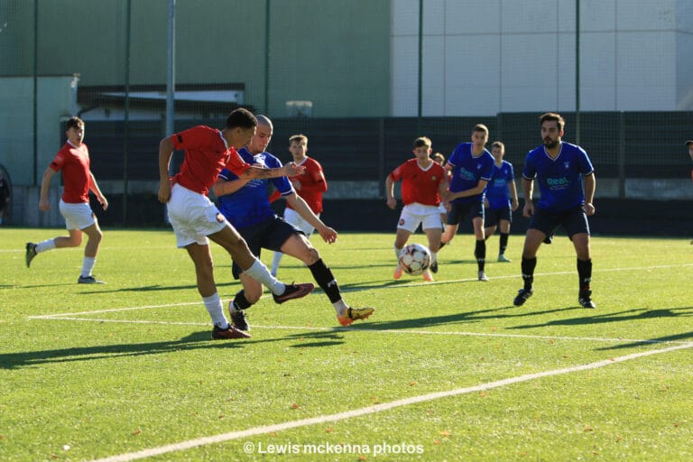 A FC United of Manchester player shoots a ball inside the Krakow Dragoons FC box, with plenty of players from both teams watching inside the box in the background