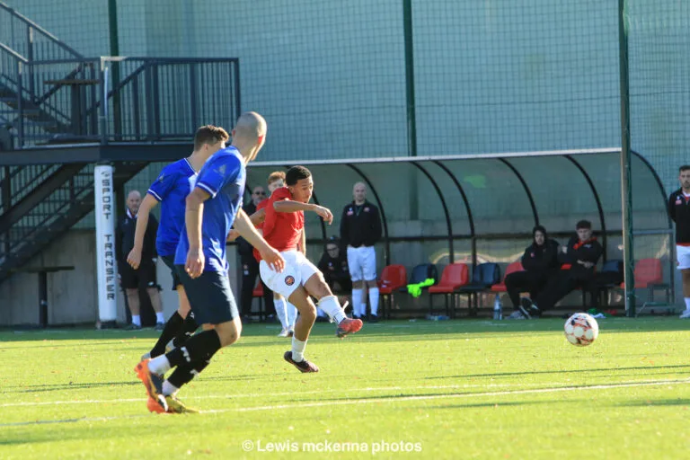 A FC United of Manchester player shoots a ball with their subs bench in the background