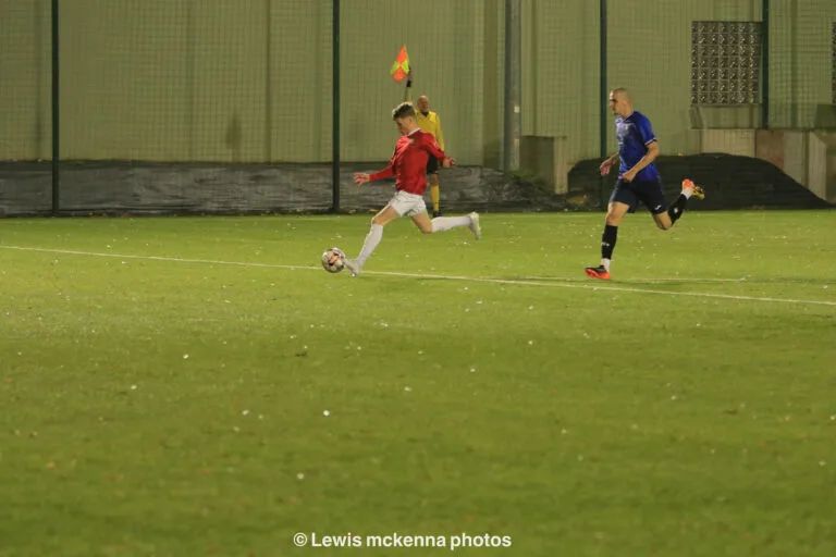 A FC United of Manchester player shooting a ball inside the Krakow Dragoons FC box, with the linesman in the background putting his flag up for offside